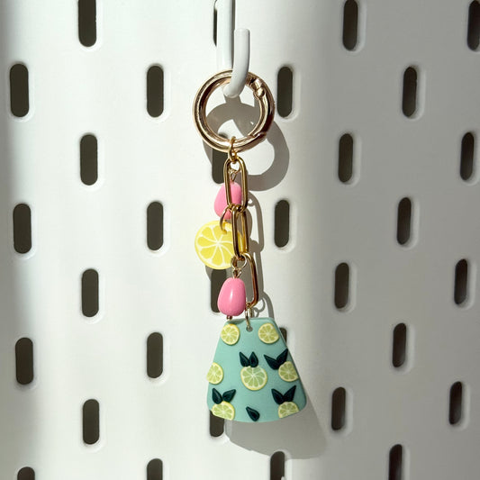 A close-up of a colorful bag charm hanging on a white perforated surface. The charm consists of a gold keyring, a gold chain with small pink bead-like accents, a yellow lemon slice charm, and a main pendant shaped like a trapezoid in pastel green, decorated with tiny lemon slices and dark green leaves. Sunlight casts a soft shadow on the background