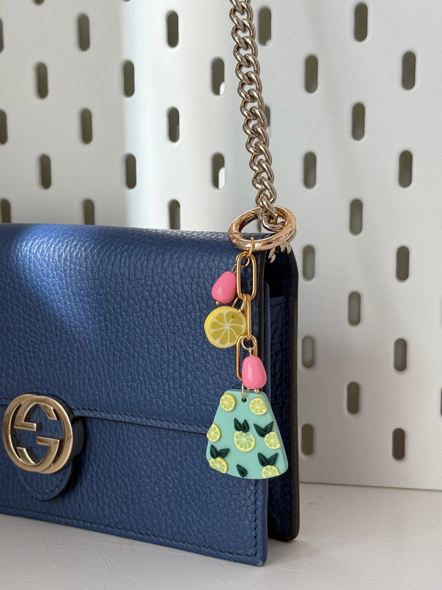 A bag charm attached to a dark blue leather Gucci bag with a gold logo and a silver chain strap. The charm dangles from the bag’s strap, showcasing its pastel green pendant with lemon slice details, pink bead-like accents, and a yellow lemon slice charm. The background features a white perforated surface. Sunlight enhances the texture of the leather and the charm's vibrant colors.