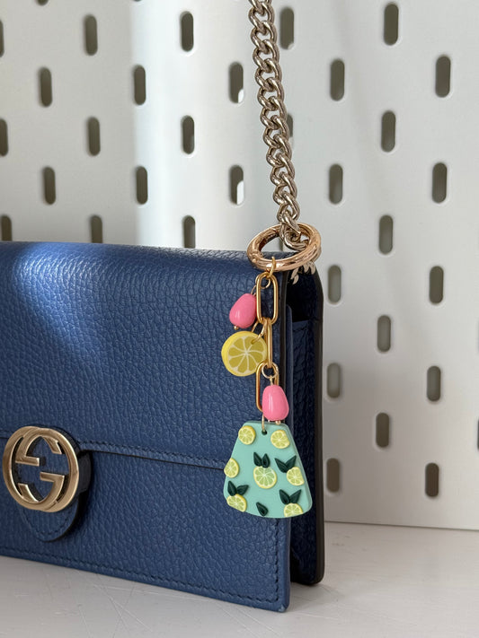  A bag charm attached to a dark blue leather Gucci bag with a gold logo and a silver chain strap. The charm dangles from the bag’s strap, showcasing its pastel green pendant with lemon slice details, pink bead-like accents, and a yellow lemon slice charm. The background features a white perforated surface. Sunlight enhances the texture of the leather and the charm's vibrant colors.